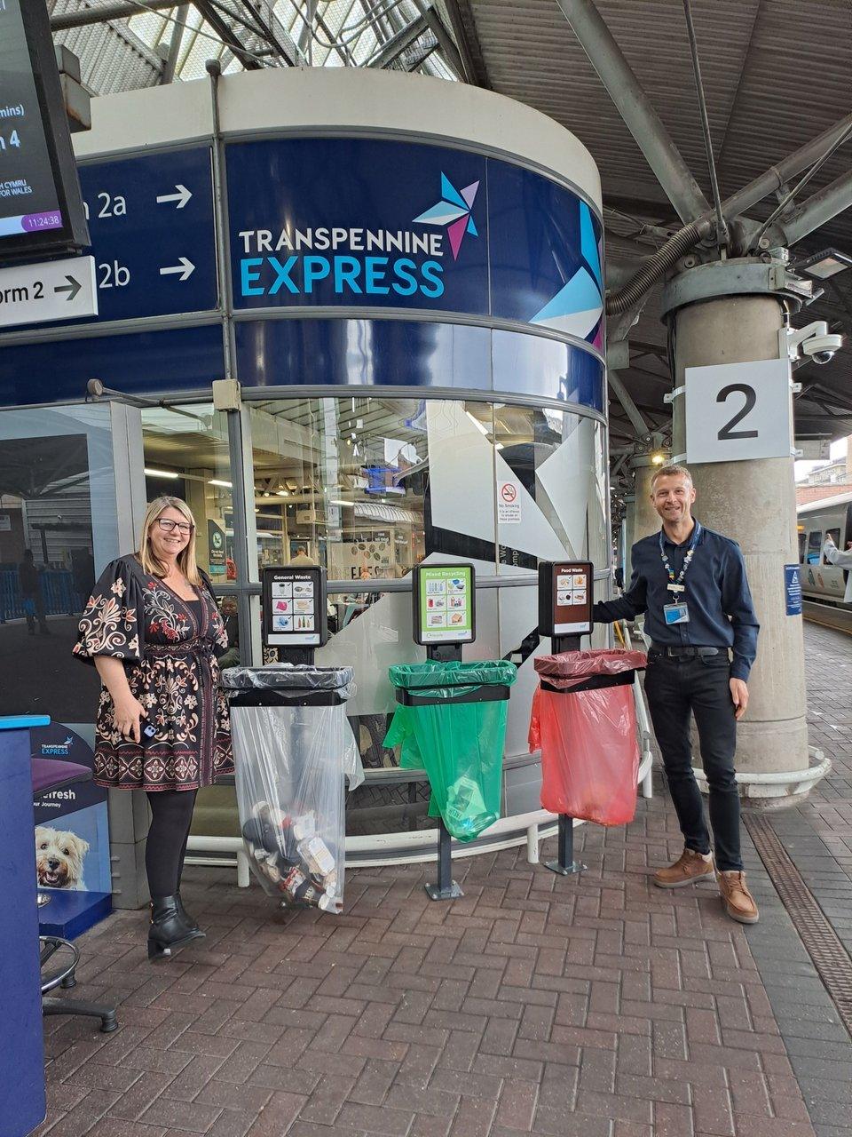Two people stand beside labelled recycling bins at a TransPennine Express station platform.
