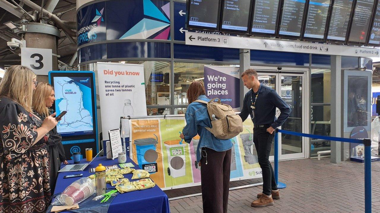 People at a recycling roadshow stand beside information displays at a train station.
