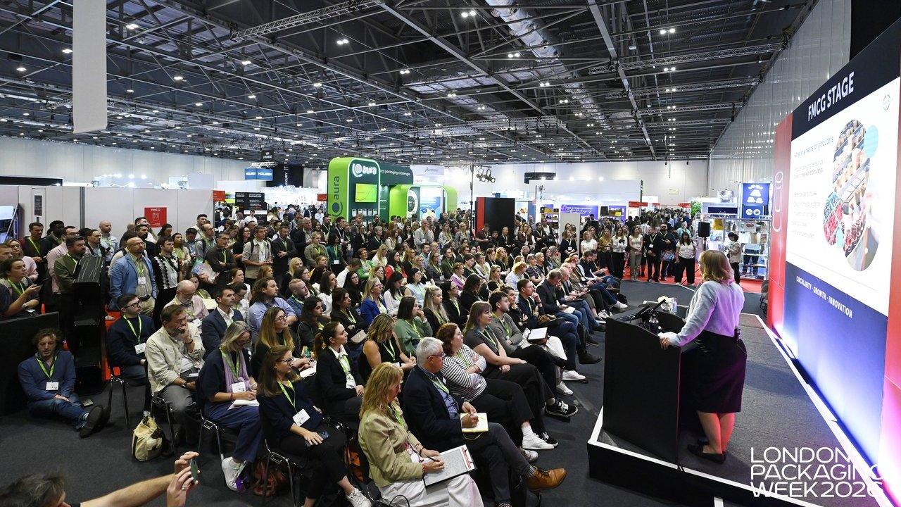 A speaker addresses a large seated audience at a packaging trade show stage.