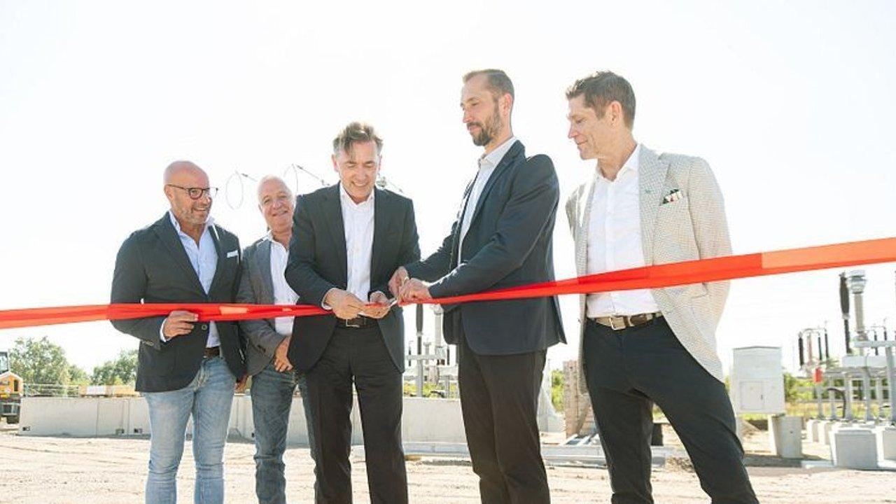 Five men in business attire cut a red ribbon at an industrial site.