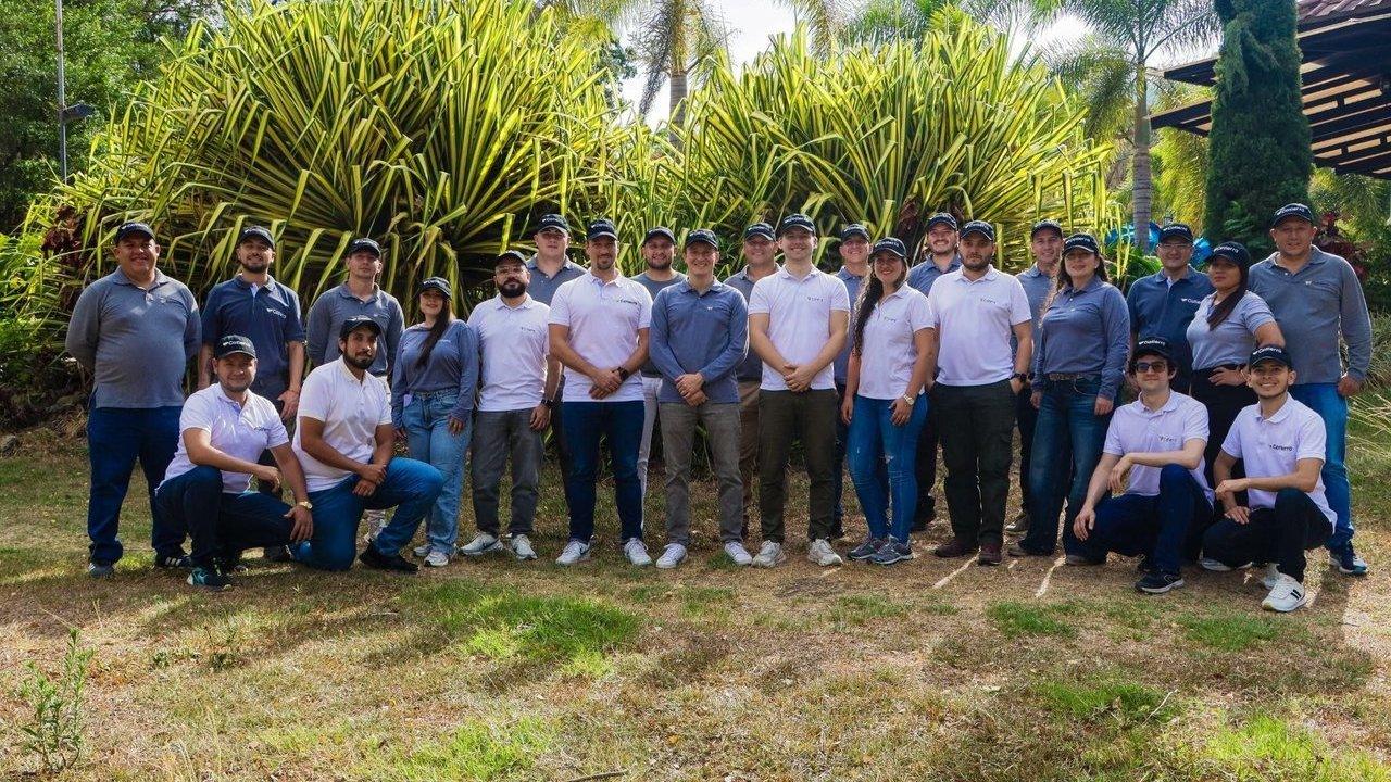 A group of 20 people in branded shirts and caps standing outdoors before tropical plants