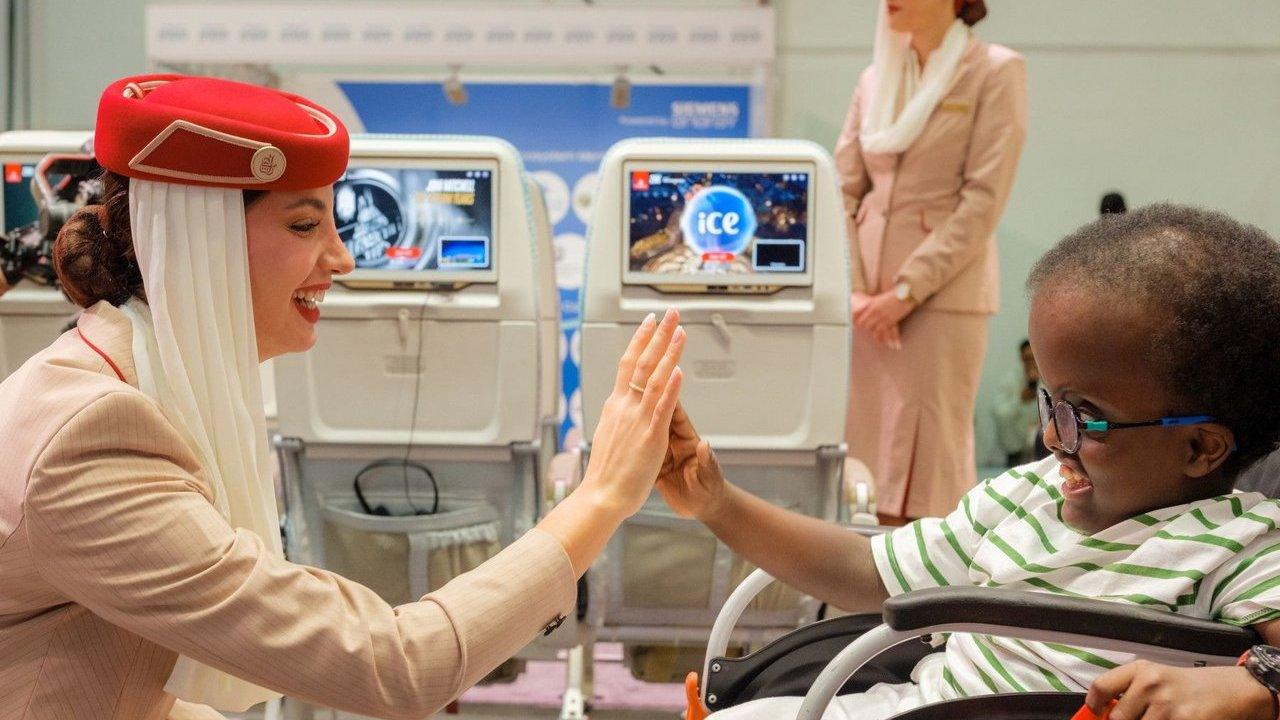 An airline attendant high-fives a smiling child in a wheelchair beside aircraft seats.