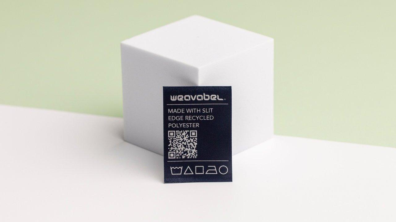 A black recycled polyester fabric label with a QR code in front of a white cube.