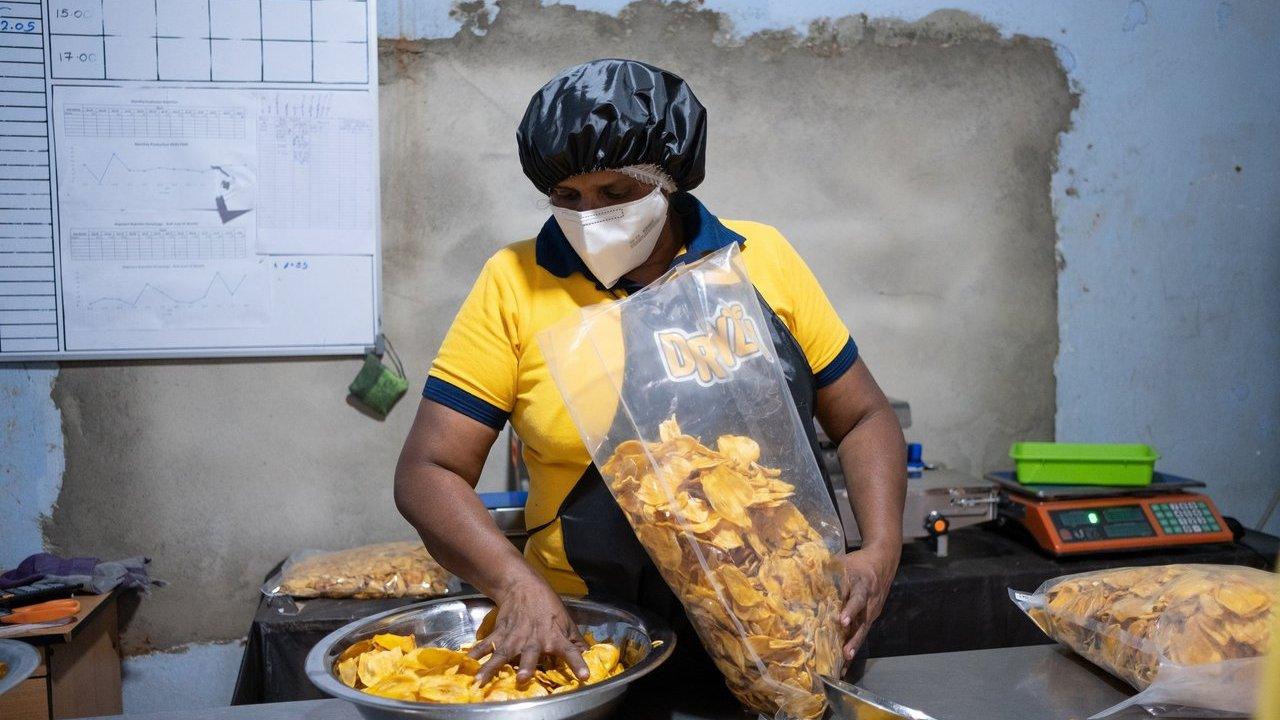 Worker in hair cover and mask packaging banana chips into a clear bag beside a scale