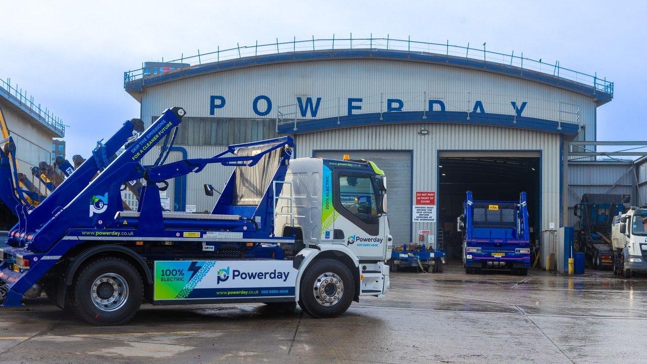 Electric skip-loader truck outside a Powerday industrial depot with other trucks by open garage doors.