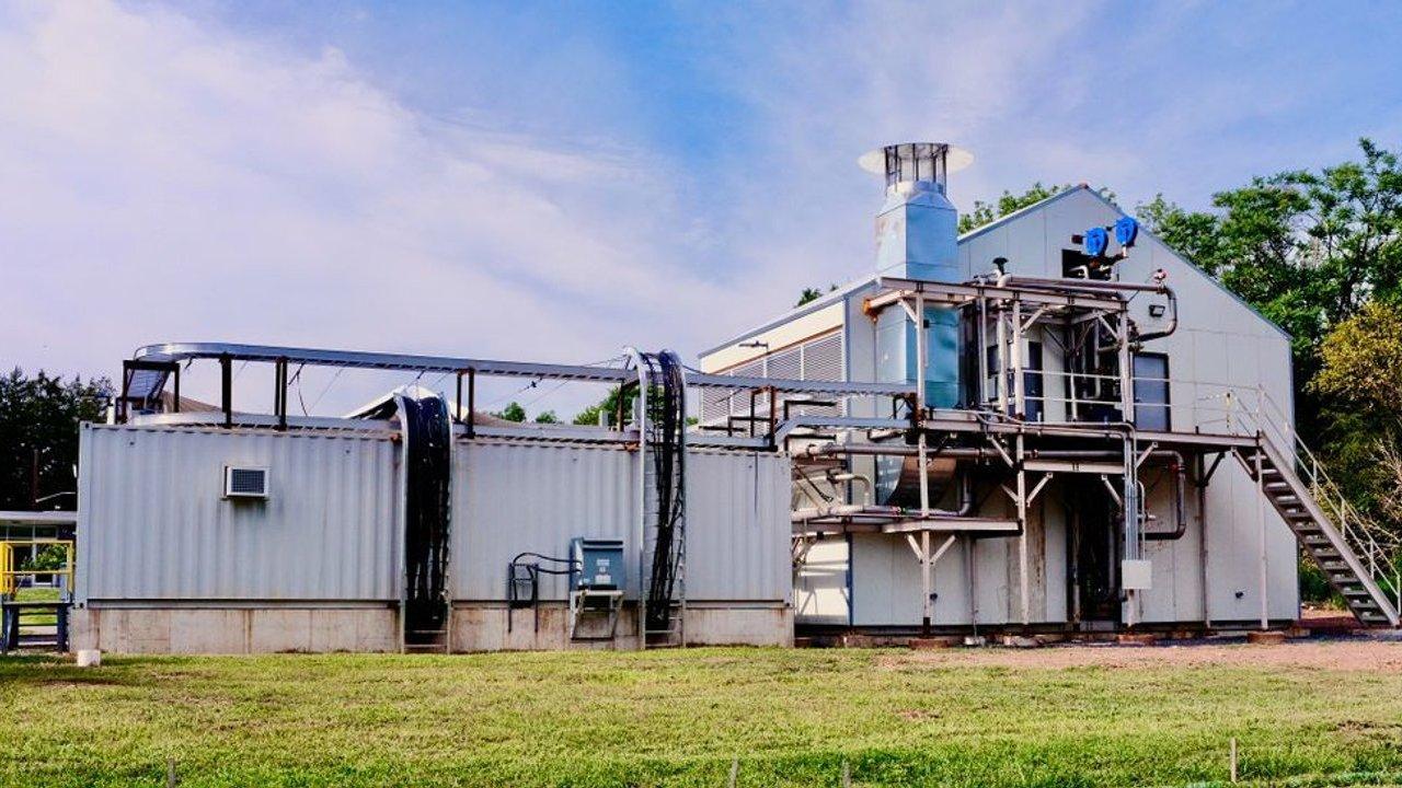 Industrial processing facility with pipes, exhaust stack and exterior metal staircase beside a grassy field.