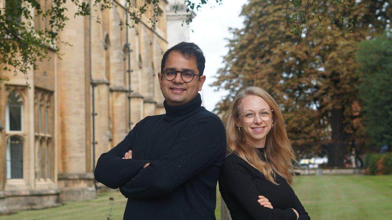 Two people standing with arms crossed outside a stone building, with trees and lawn behind them.