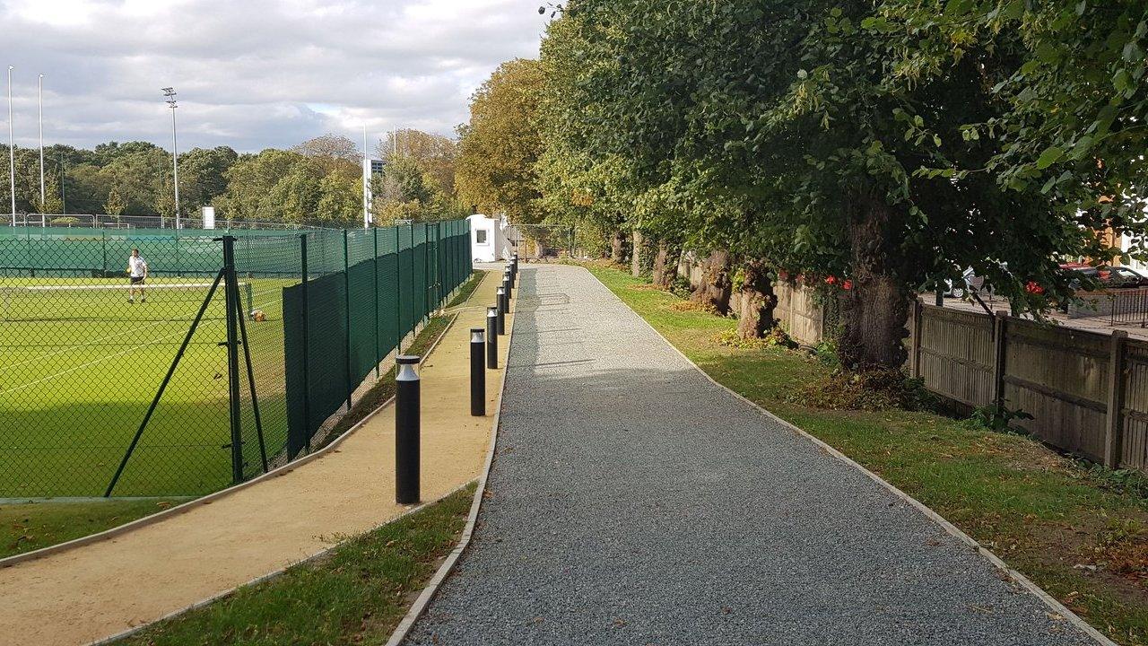 Gravel footpath beside a fenced sports field, lined with bollards and trees.