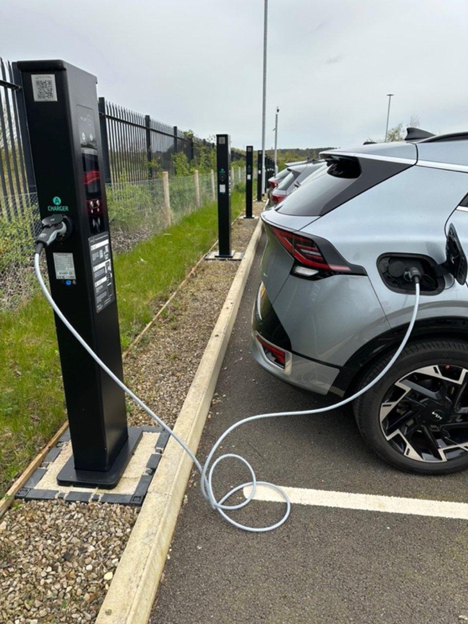 Silver electric car charging at a row of black charging stations beside a fence.