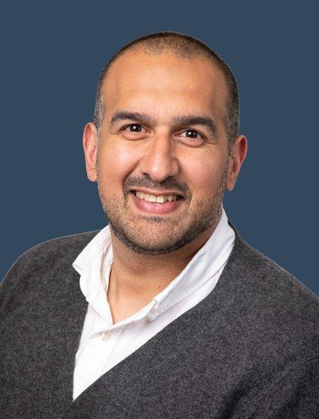 Amman Boughan Pagabo smiling in a white shirt and gray sweater against a blue background