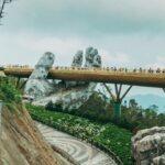A golden pedestrian bridge crosses giant stone hands on a forested mountainside.