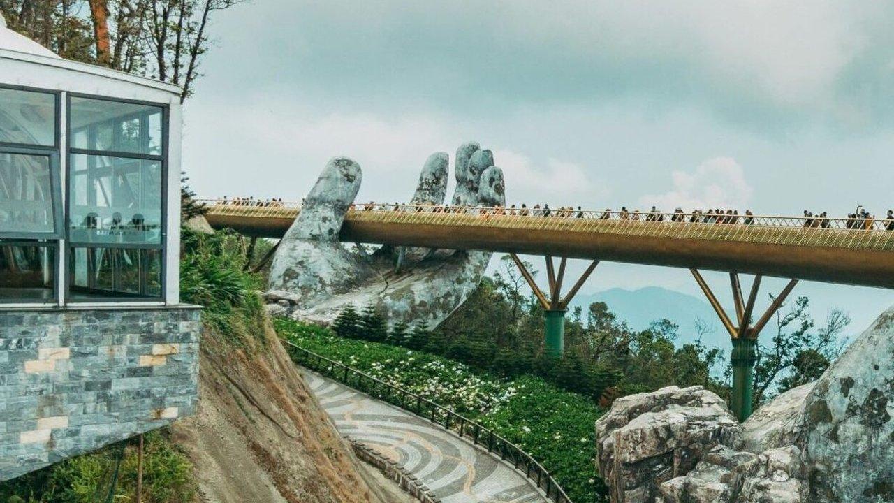 A golden pedestrian bridge crosses giant stone hands on a forested mountainside.