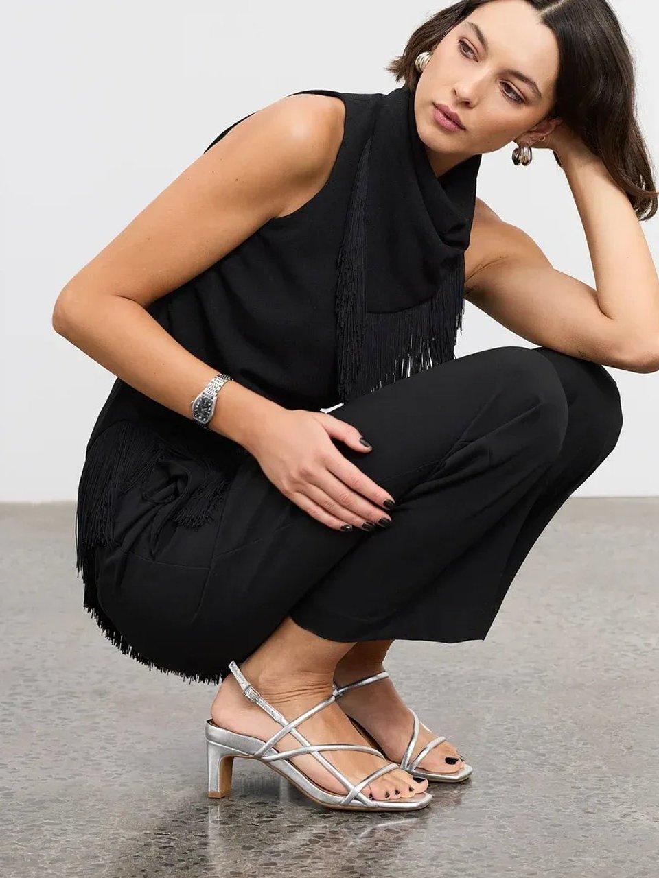 Woman in black outfit wearing silver strappy heeled sandals while crouching.