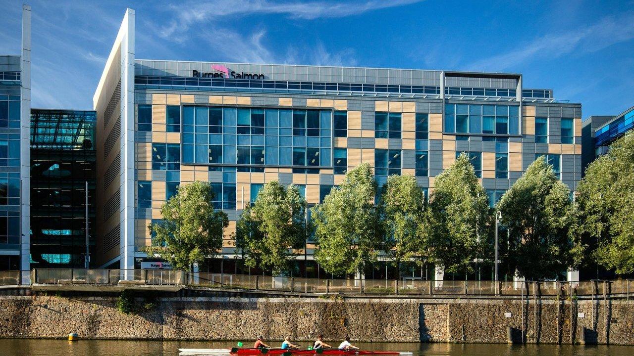 Burgess Salmon office building beside a river, with trees along the waterfront and rowers in a boat.