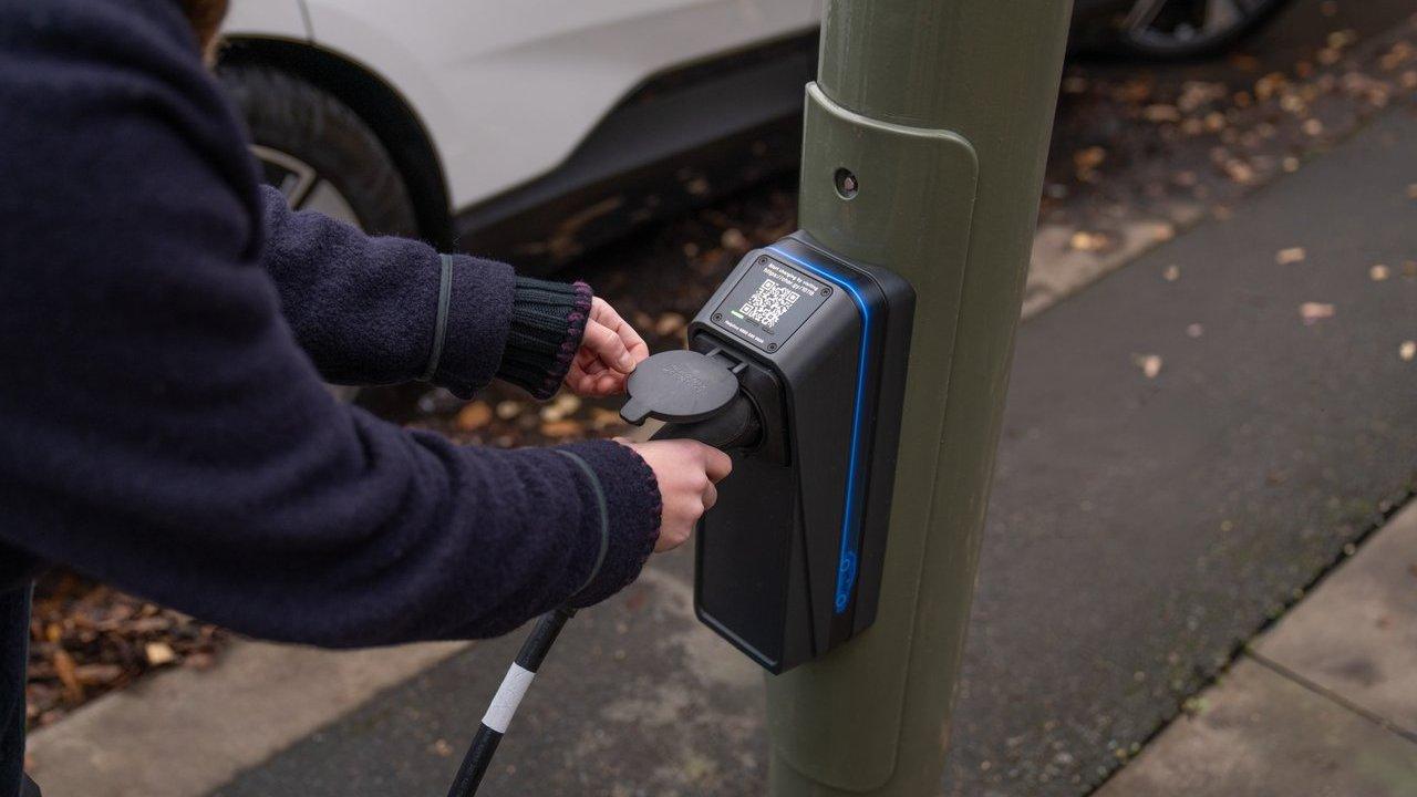 Person plugging an electric vehicle charging cable into a curbside charger mounted on a pole.