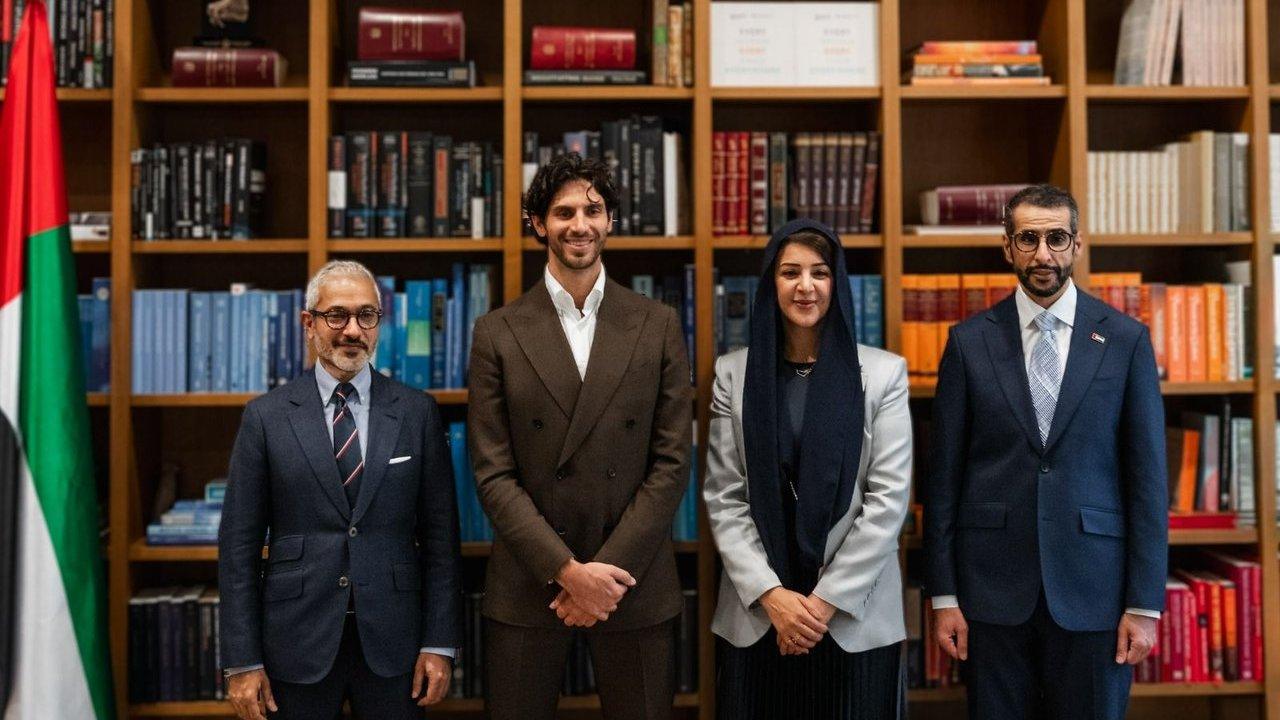 Four people in formal attire stand before bookshelves beside a United Arab Emirates flag.
