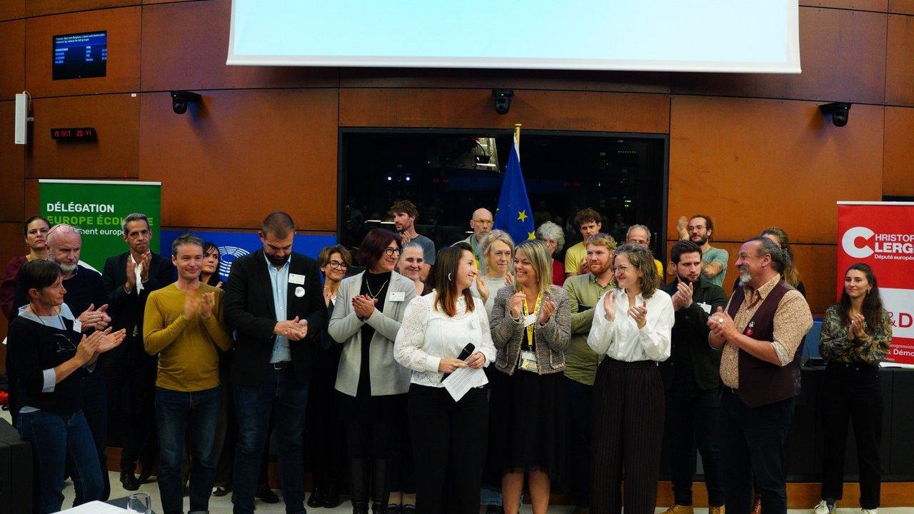 A group of people applauding around a woman holding a microphone, with European Parliament banners and an EU flag behind them