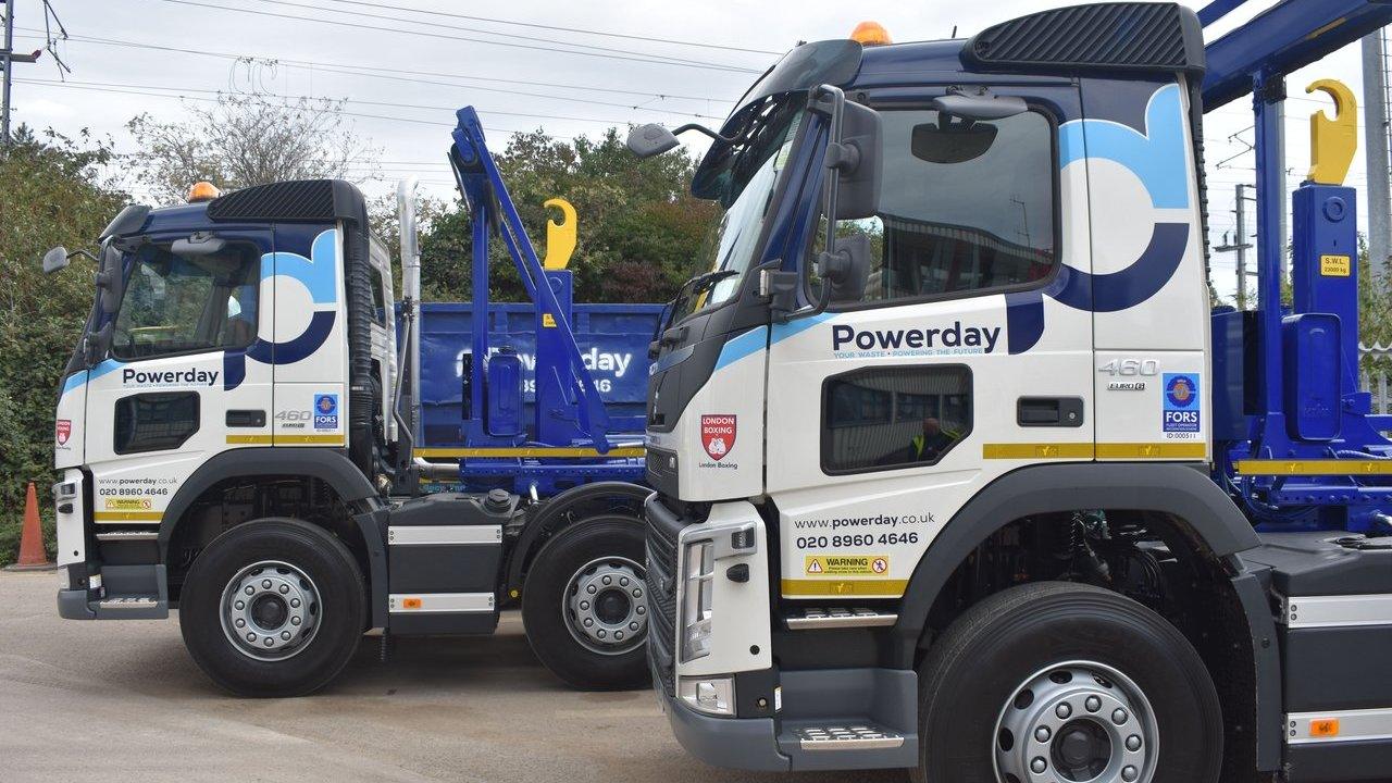 Two blue-and-white Pow­erday hook-lift trucks parked side by side.