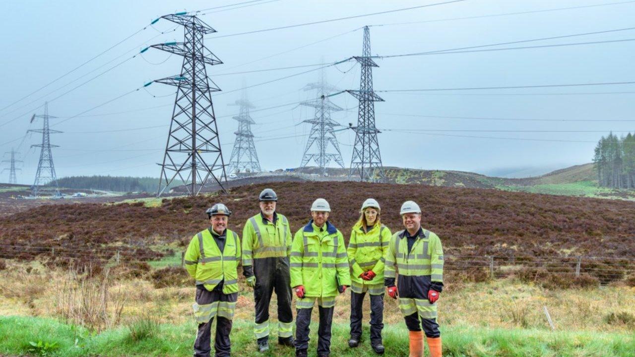Five utility workers in high-visibility clothing stand beneath electricity pylons in a rural landscape.