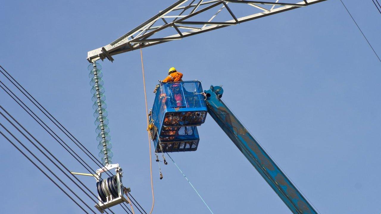 Utility worker in a raised basket repairs high-voltage power lines beneath a transmission tower.