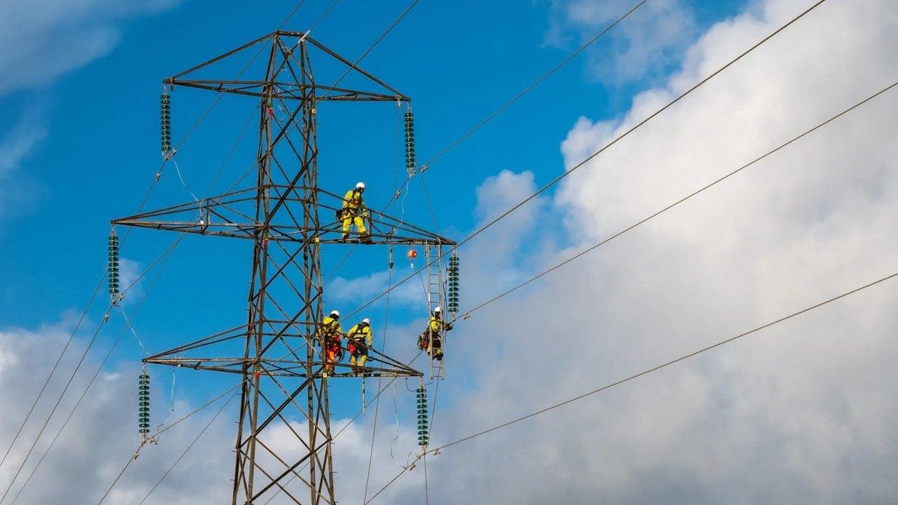 Utility workers in yellow safety gear on a high-voltage electricity pylon under a blue sky.