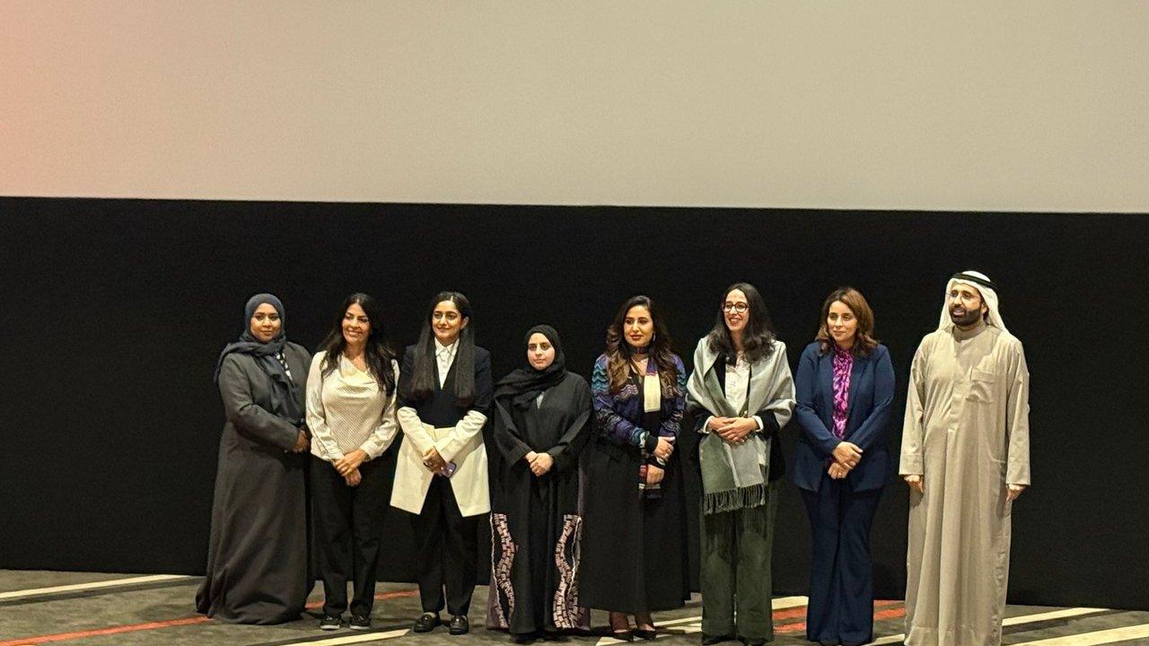 Eight people standing in a row against a black wall, including women in formal attire and a man in traditional dress