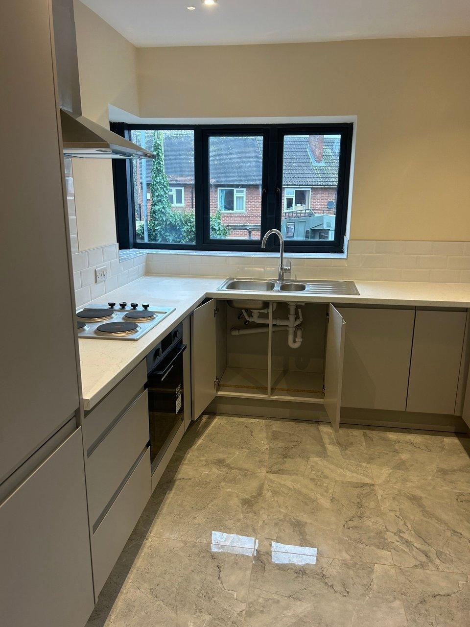 Kitchen with gray cabinets, electric hob, oven, double sink and open cabinet beneath a large window.