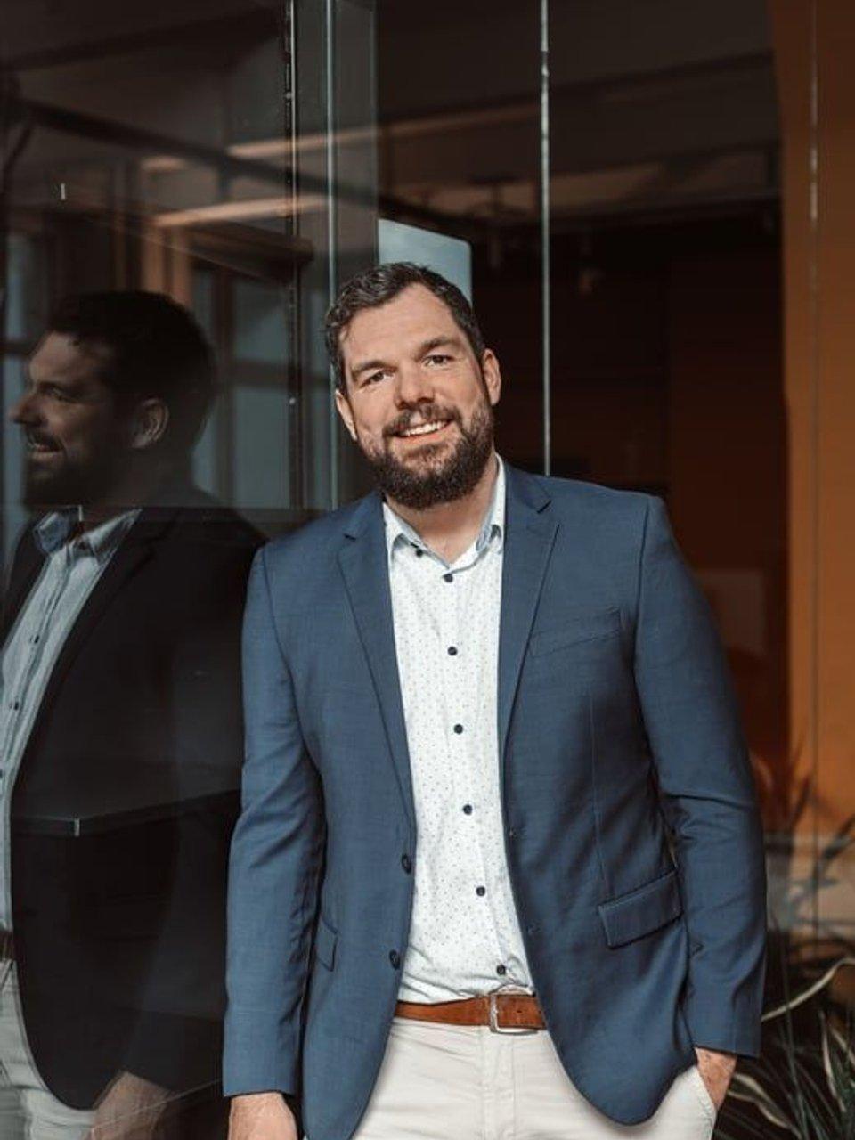 Jan Steggewentz in a blue blazer standing beside a glass wall with his reflection visible.