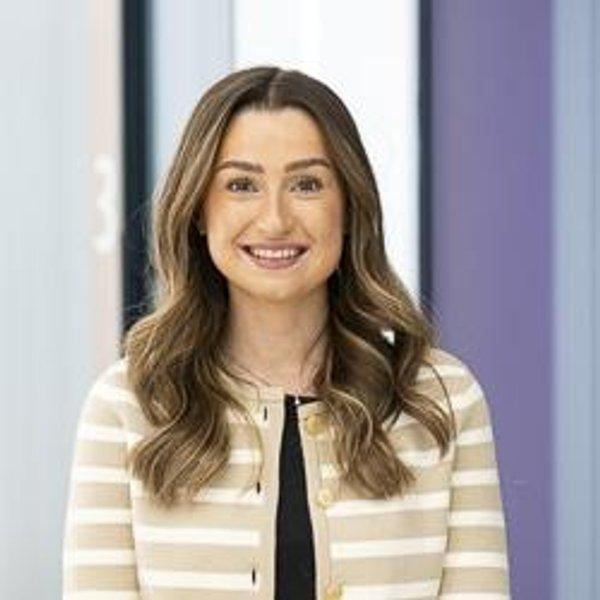 Smiling woman with long brown hair wearing a beige striped jacket.