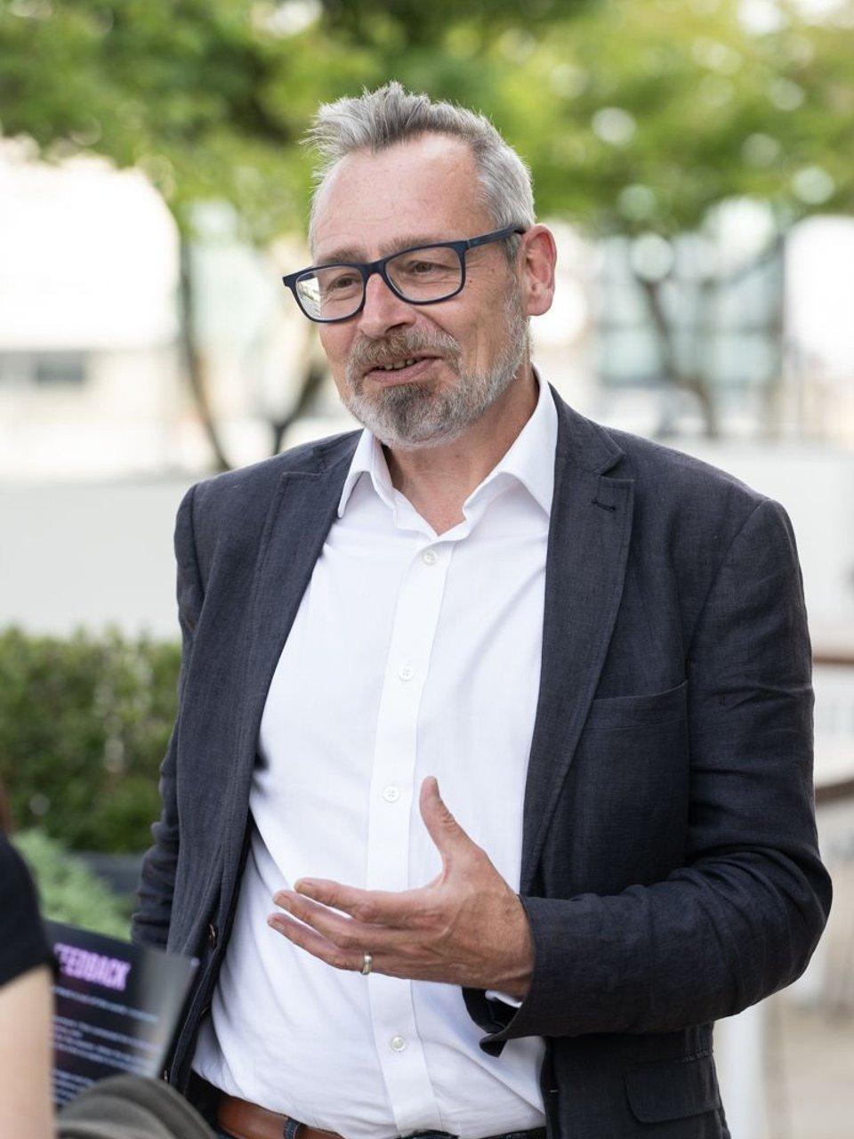 John Garner in a dark blazer and glasses speaking outdoors.