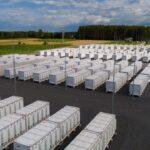 Rows of white battery storage containers at an energy facility beside fields and woodland.