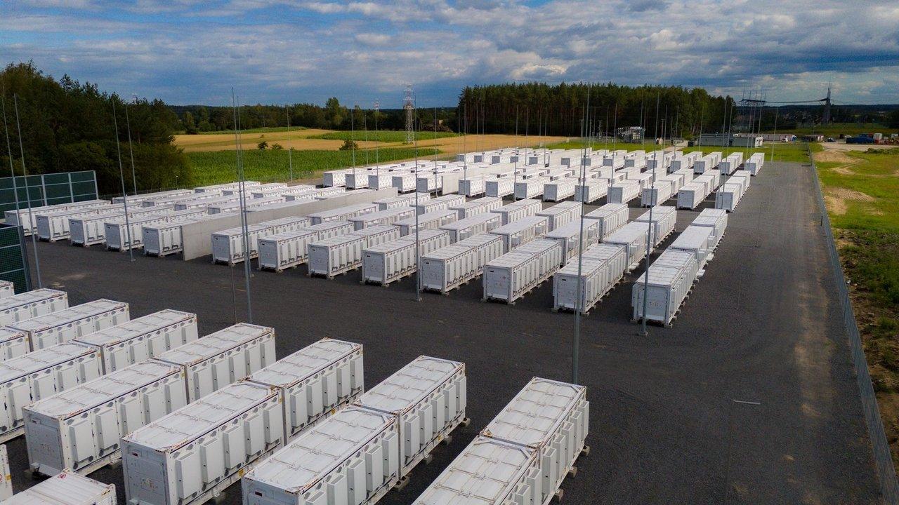 Rows of white battery storage containers at an energy facility beside fields and woodland.