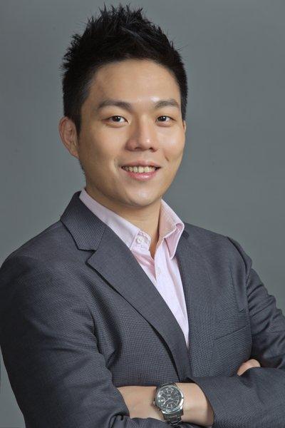 Melton Chang in a gray suit and pink shirt, smiling with arms crossed against a gray background