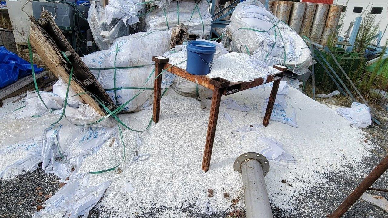 Spilled white plastic pellets surround torn bulk bags, a rusty table and a blue bucket.