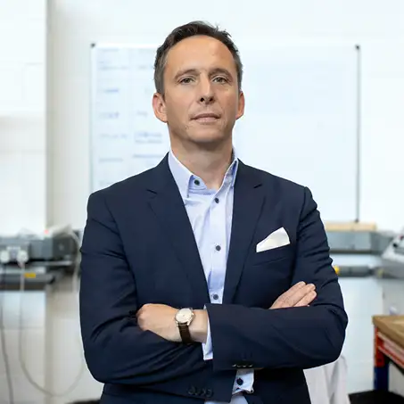 Man in a dark suit standing with arms crossed indoors.