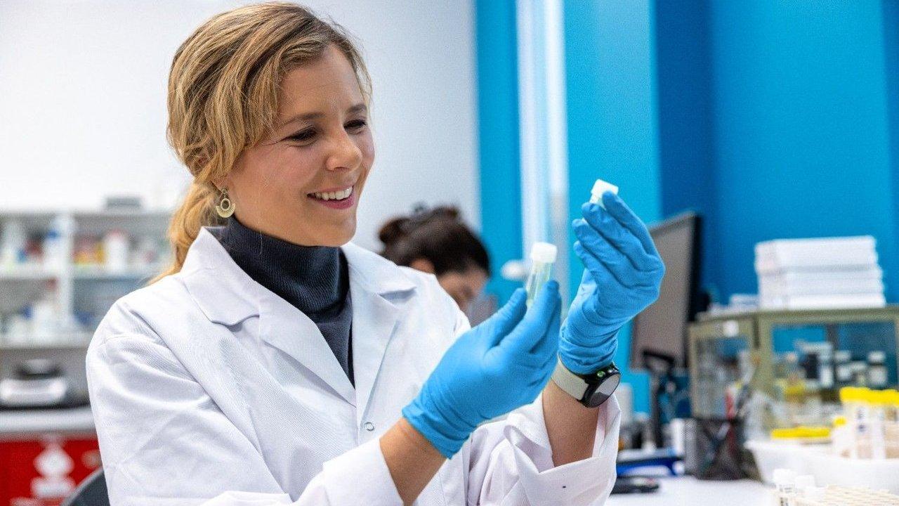 Kate Jones in a lab coat and blue gloves examines a small vial in a laboratory.
