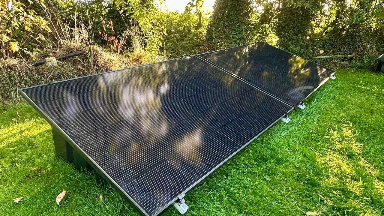 Three solar panels mounted low on a grassy lawn beside dense trees and shrubs.