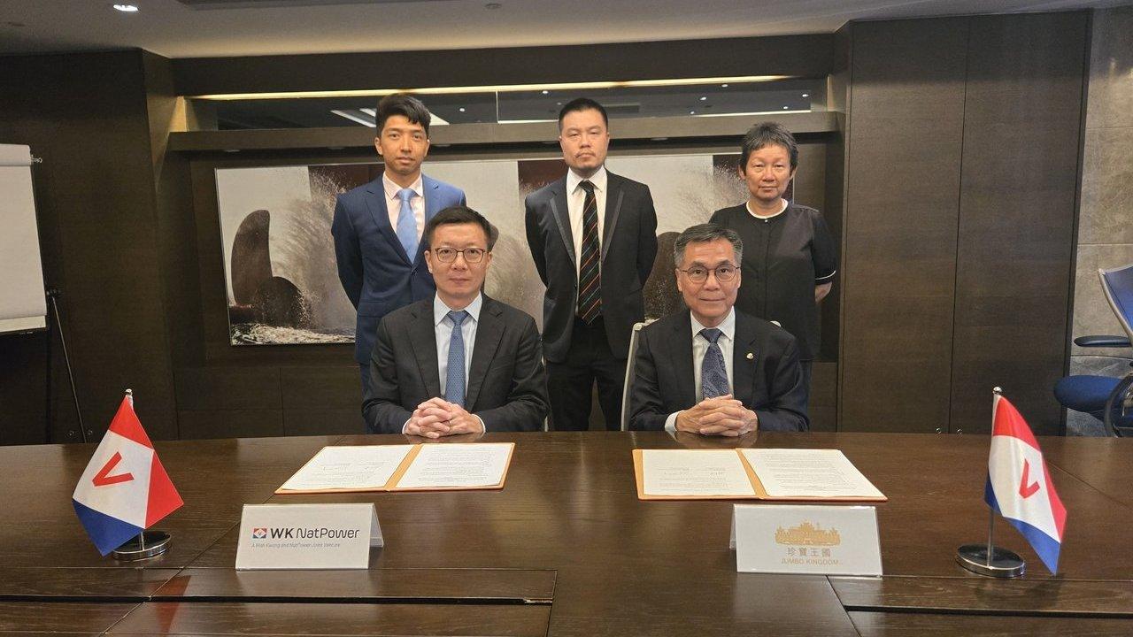 Two executives seated at a signing table with documents, flags and four colleagues standing behind them.