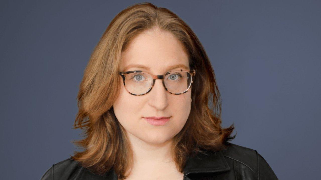 Rebecca Brocato wearing glasses and a black jacket against a blue-gray background.