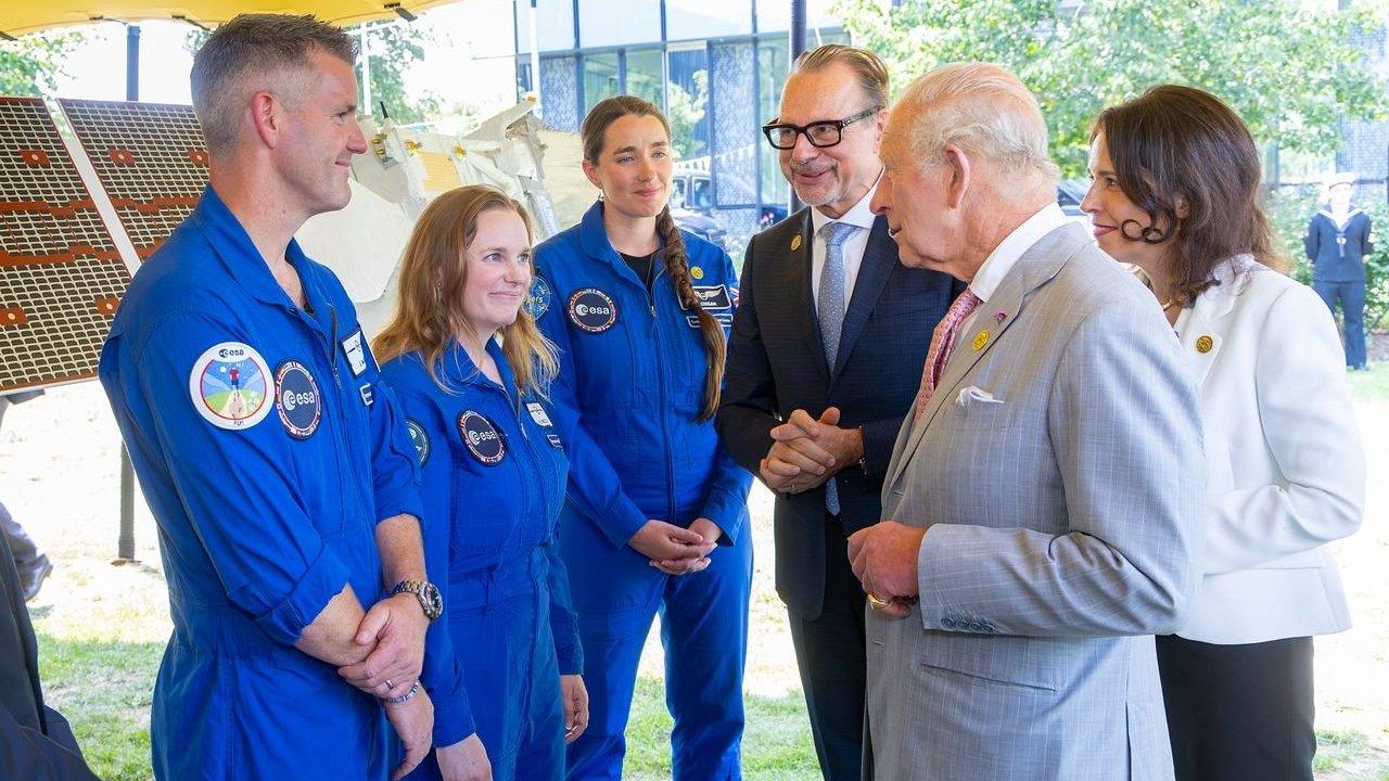 HM King Charles III speaks with European Space Agency astronauts in blue flight suits beside a satellite display.