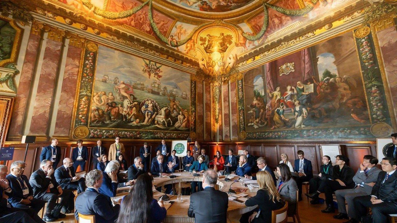 Business leaders seated around a conference table in an ornate frescoed hall.