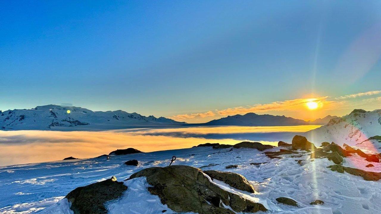 Sunrise over snow-covered mountains and a sea of clouds, with rocks in the foreground.