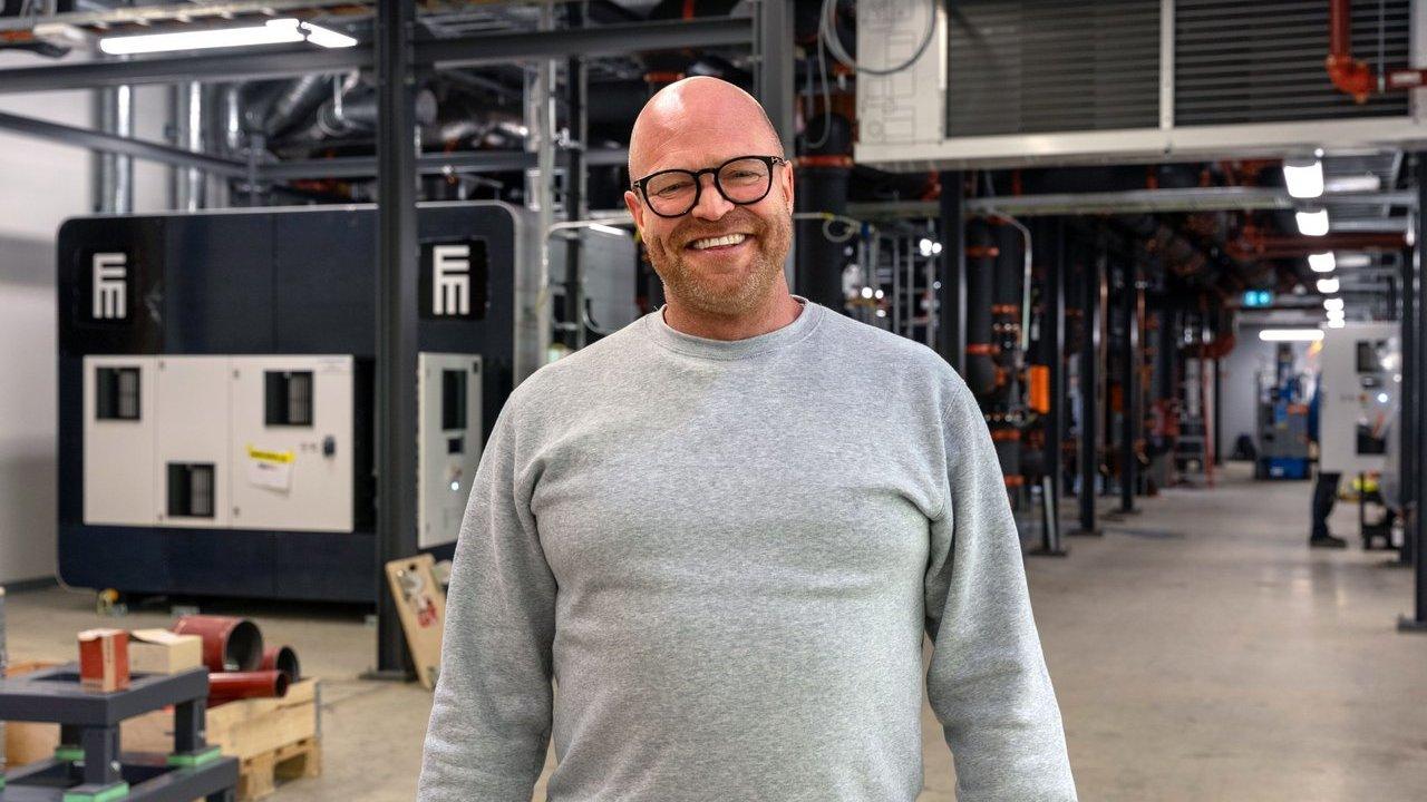 A man in glasses stands in an industrial facility with pipes and equipment.