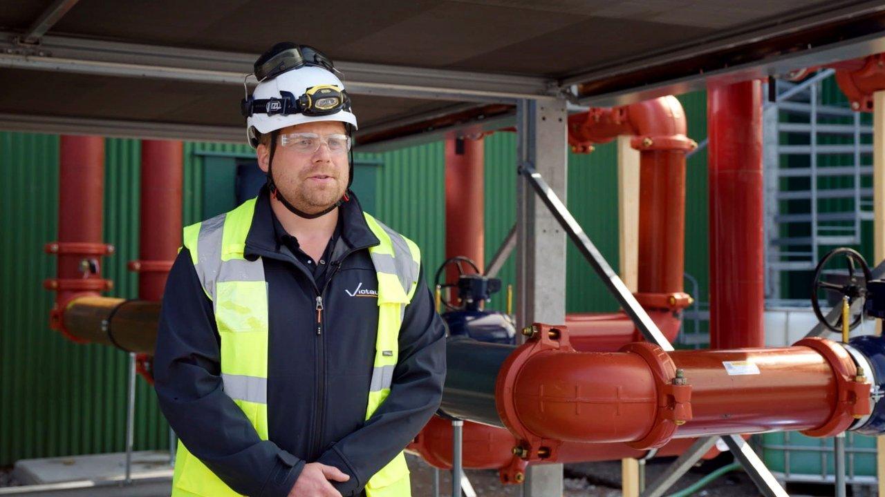 Worker in high-visibility vest and hard hat standing beside red industrial pipes and valves