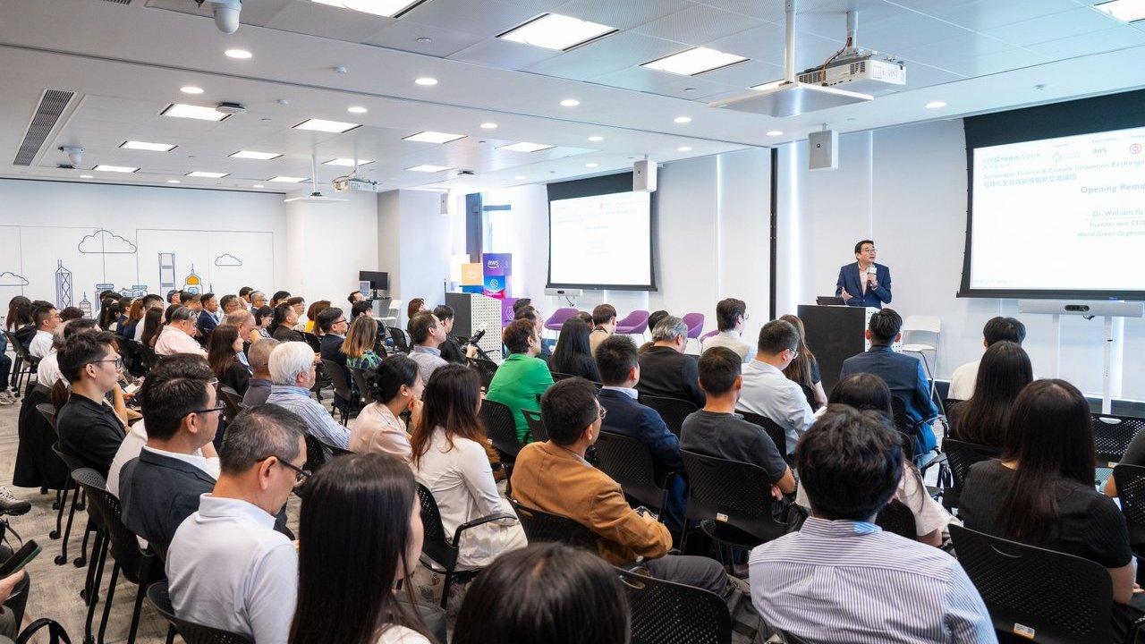 Audience seated in a conference room listening to a speaker beside projected presentation slides.
