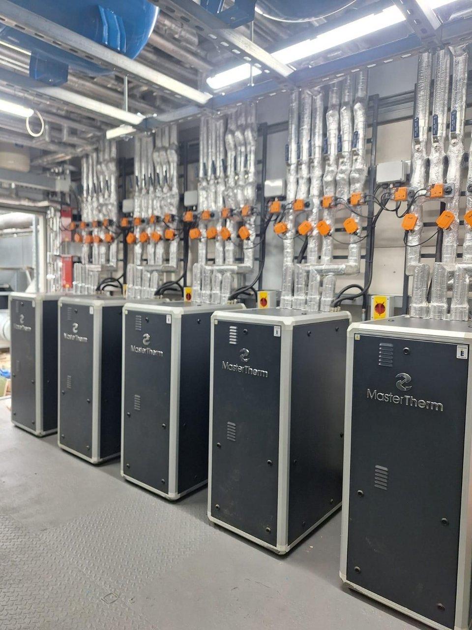 Five heat pump units beside insulated pipework and orange control valves in a plant room.