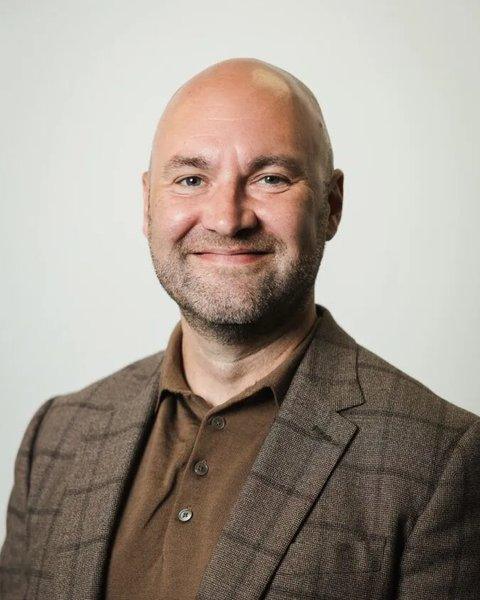 A smiling bald man in a brown shirt and checked blazer against a light background.
