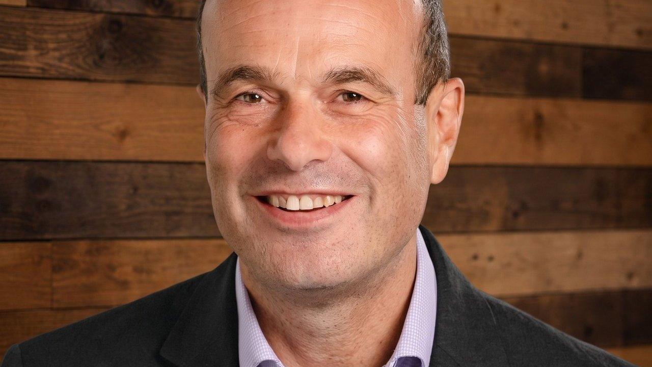 Smiling man in a dark jacket and light shirt against a wooden plank wall