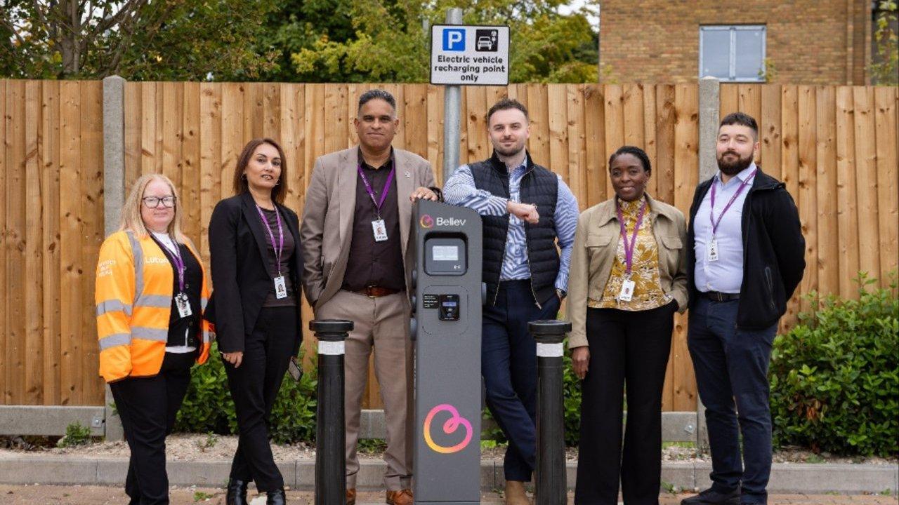 Six people stand beside a Believ electric vehicle charging point beneath a parking sign.