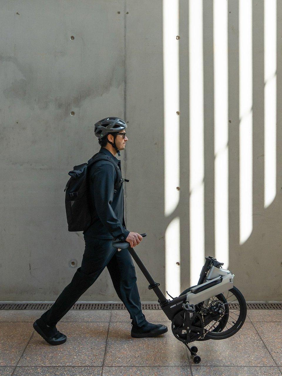 Man wearing a helmet and backpack walks beside a folded electric bicycle.